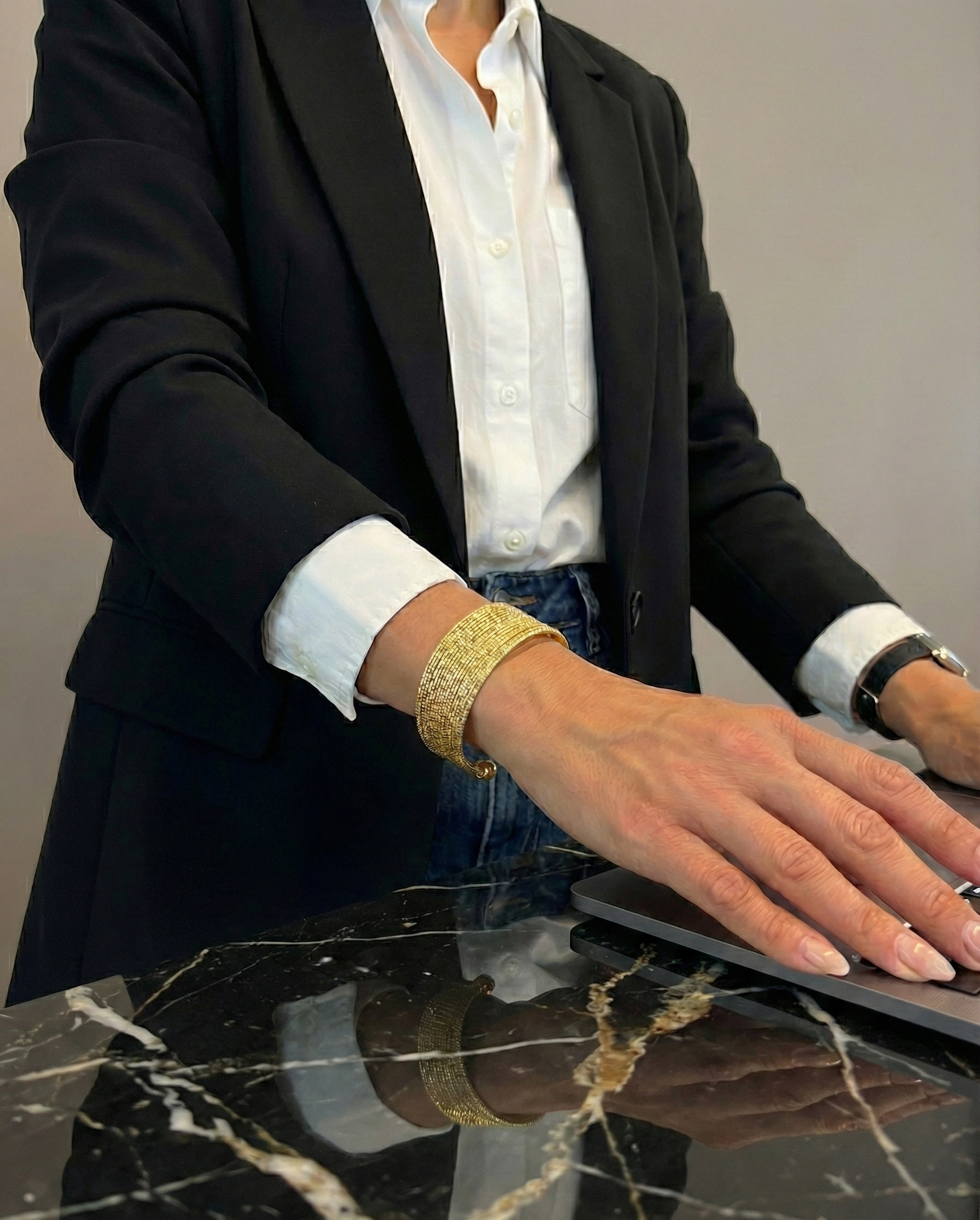 Person wearing a black blazer and white shirt with a gold bracelet on a marble surface.